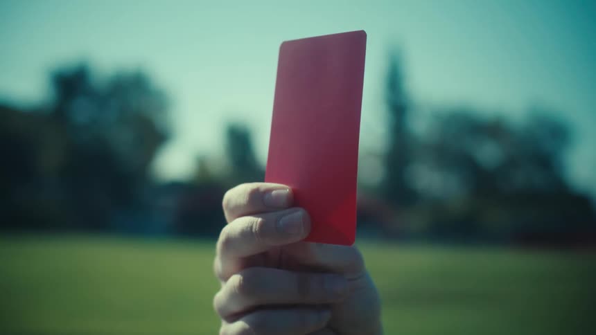 A football referee raises a red card in slow motion, issuing a serious penalty on a green pitch