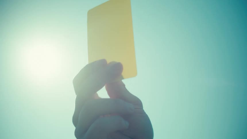 A referee's hand holds up a yellow penalty card against the bright sun and blue sky in slow motion