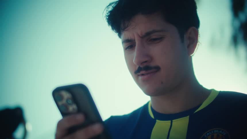 A young man with a mustache wearing a soccer jersey looks down and scrolls on his smartphone on a bright sunny day