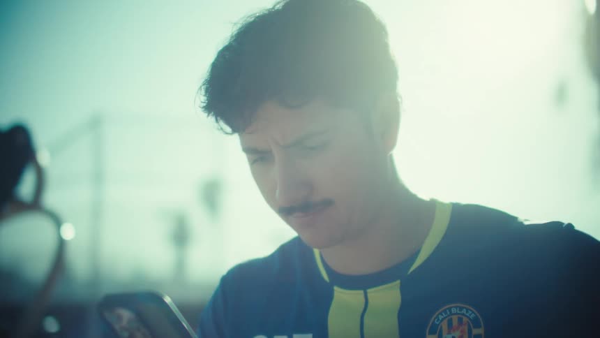 A young man in a soccer jersey looks at his phone with a serious expression while standing in a sun-drenched park