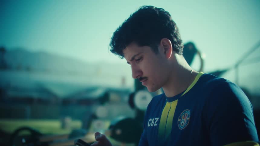 A young male soccer player in uniform sits on an outdoor exercise bench and smiles while using his smartphone