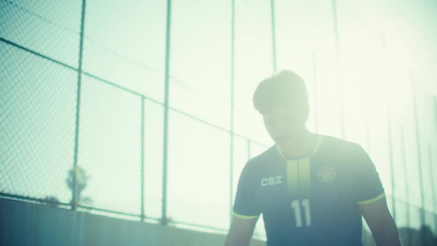 A young soccer player juggles a ball in slow motion while walking forward on a sun-bleached street