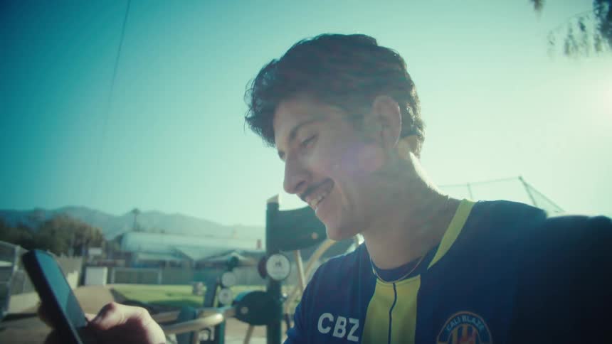 A young male soccer player in his uniform relaxes at a sunny outdoor gym, smiling happily as he texts on his smartphone