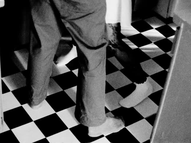 A couple's feet slow dance across a classic black and white checkerboard floor in grainy, vintage-style footage
