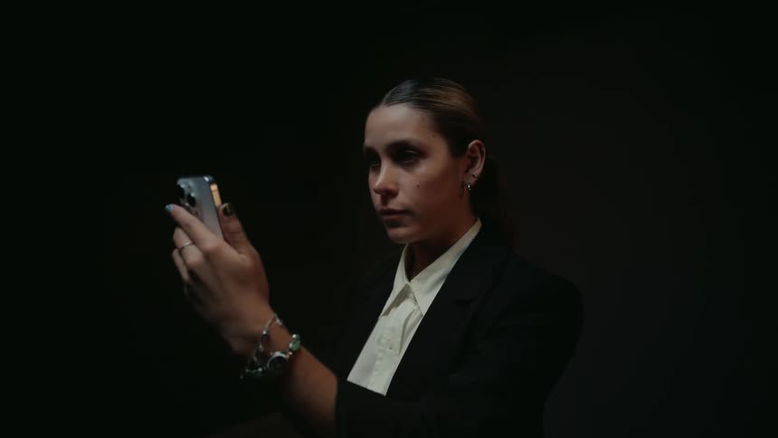 A serious woman in a blazer uses her smartphone in a dark room, her face illuminated by the screen's glow
