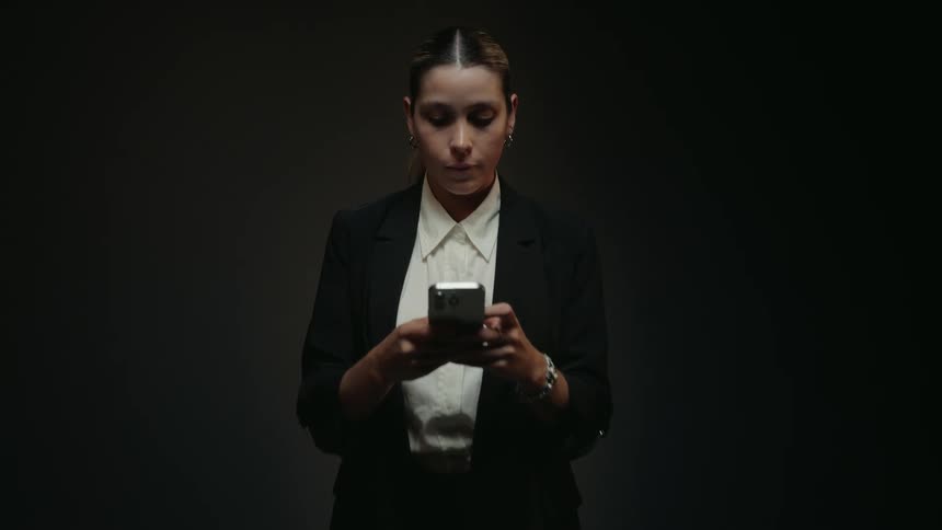 A serious businesswoman stands alone in a dark, spotlit room, intently focused on her smartphone as the camera slowly zooms in