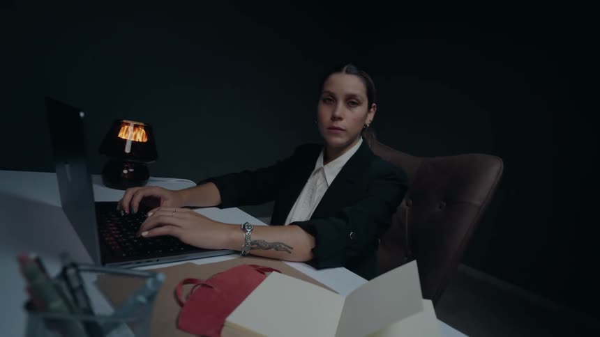 A serious businesswoman in a dark suit works on her laptop in a dimly lit office before looking directly at the camera