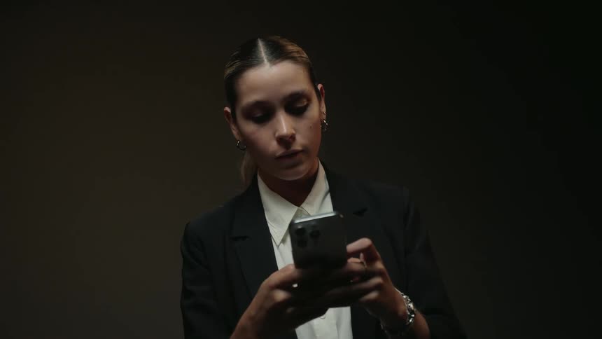 A focused businesswoman in a suit uses her smartphone in a dimly lit room with dramatic lighting