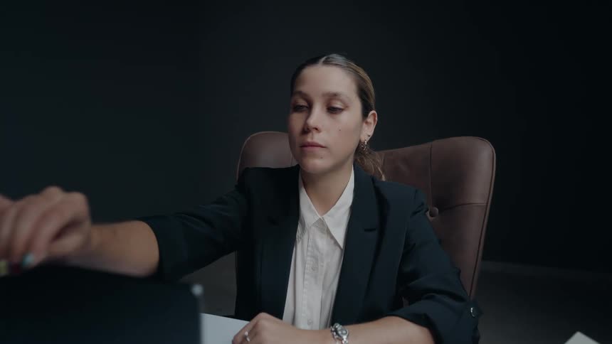 A focused businesswoman in a dark room prepares for work by slowly opening her laptop on the desk