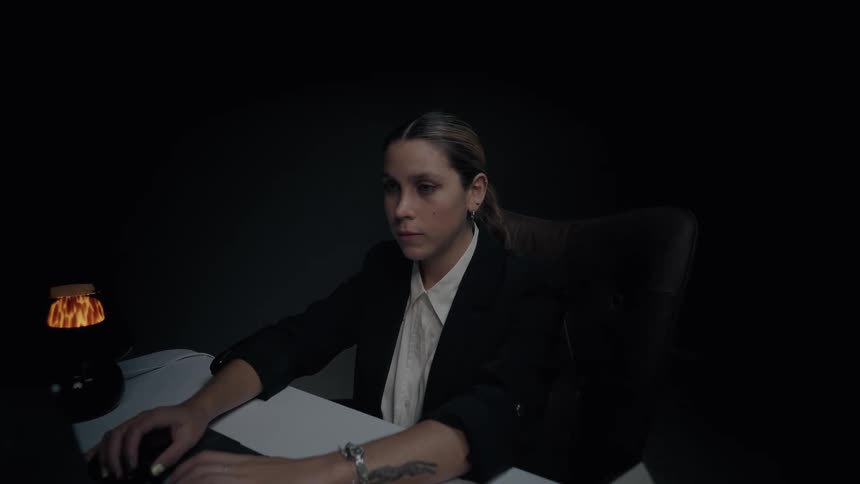 A focused businesswoman in a dark suit works late at night on her laptop in a dimly lit office before looking up and smiling at the camera