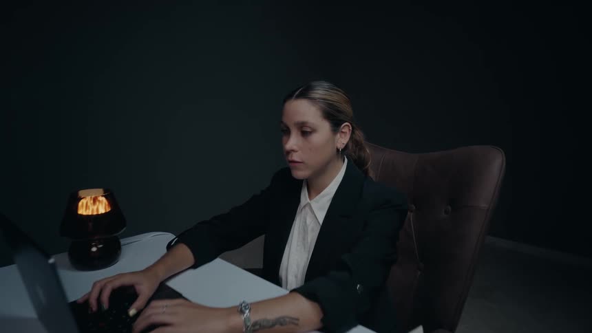 A determined businesswoman types on her laptop in a dark, minimalist office, then looks up to smile confidently at the camera