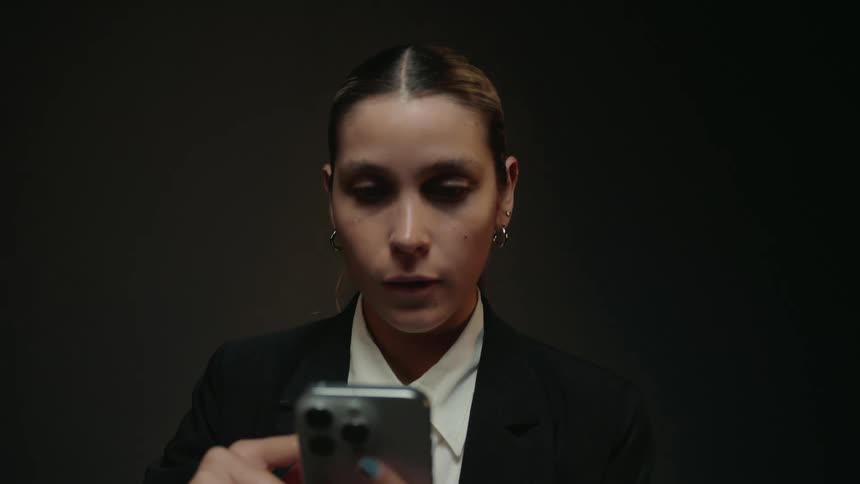 A focused businesswoman in a black suit scrolls on her smartphone in a dimly lit studio