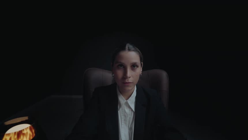 A businesswoman sits at a desk in a dark room, finishes working on her laptop, and looks intently at the camera with a slight smile