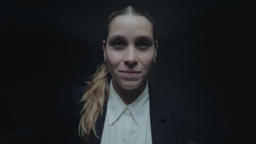Close-up of a woman in business attire whose blank expression slowly turns into a sinister smile in a dark room