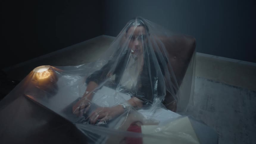 A stressed woman works on a laptop in a dark room, trapped under a large plastic sheet, symbolizing burnout and isolation