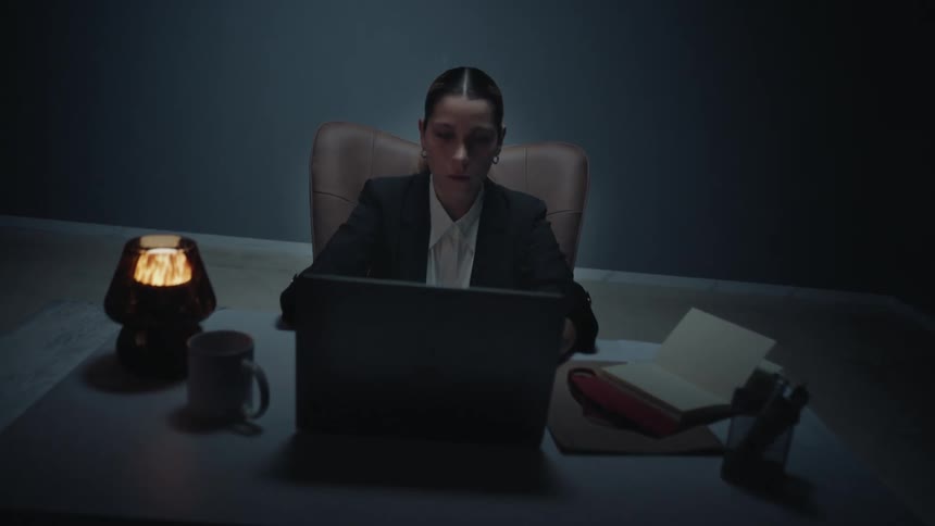 Dolly in shot of a stressed businesswoman working late in a dark office, stopping to clutch her head in pain from a headache