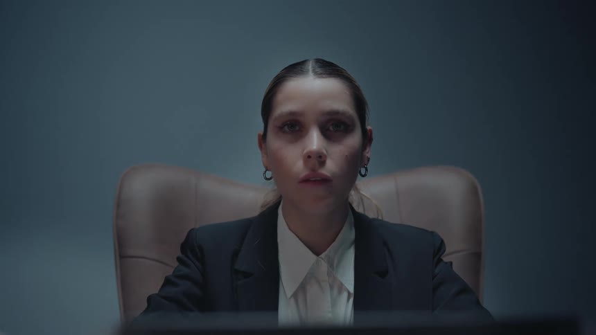 A serious businesswoman in a suit stares intently while working on a laptop in a dimly lit office as the camera slowly pushes in on her face