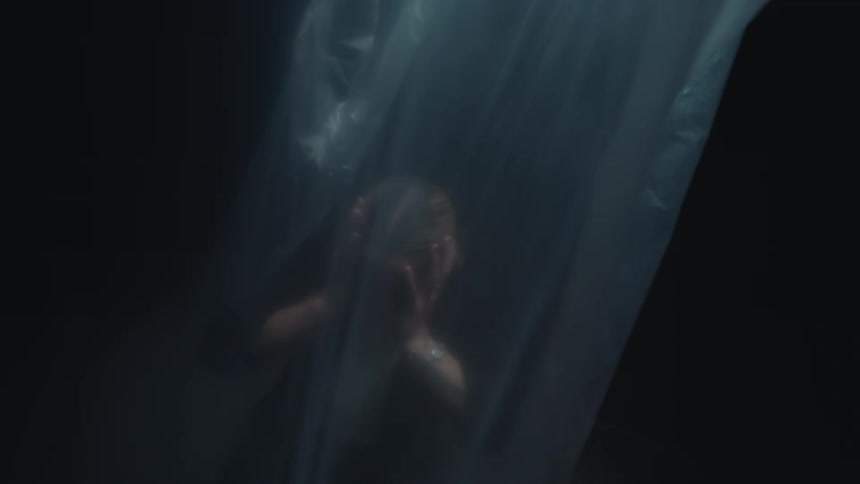 A terrified woman in a dark, hazy room covers her ears in distress behind a translucent sheet