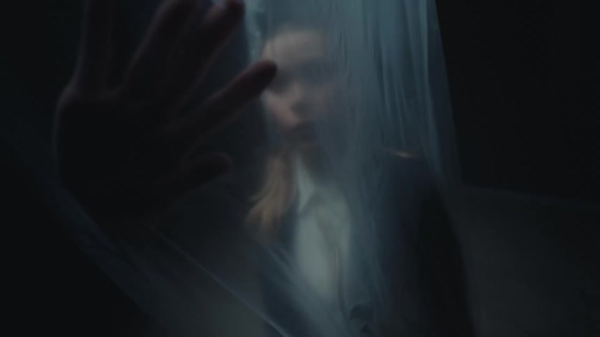 High-angle slow-motion shot of a woman in a suit trapped behind a plastic sheet, reaching her hand out towards the camera in a dark room