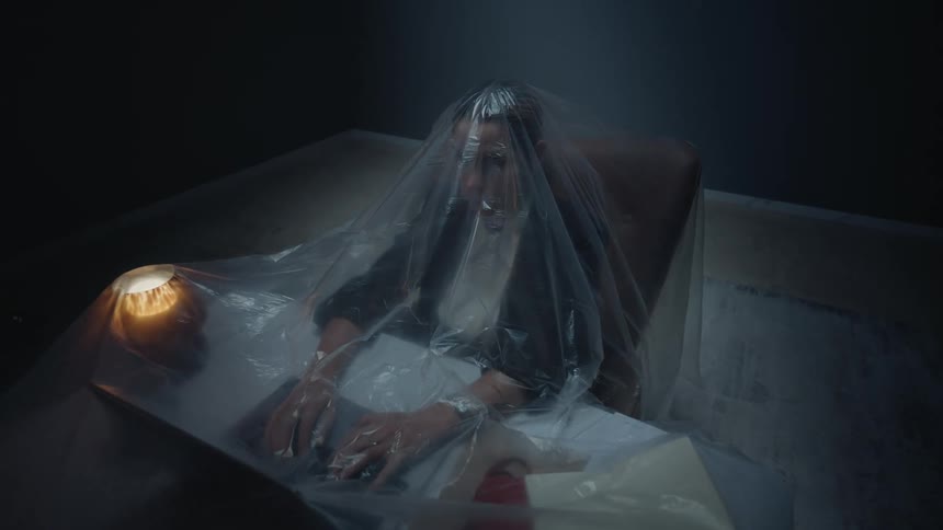 High-angle dolly-in shot of a woman in a dark room trapped under a plastic sheet while working at her desk