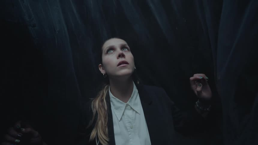 A fearful young woman in a suit looks up in a dark, plastic-wrapped room as mysterious hands rise around her