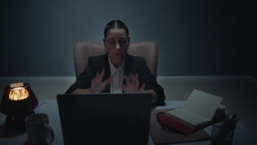 Dolly-in shot of a stressed and overworked businesswoman sitting at her desk in a dark room, holding her head in exhaustion