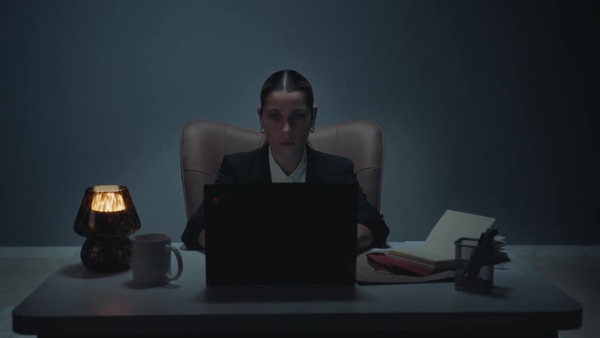 A stressed businesswoman works late on her laptop in a dark office, showing signs of exhaustion as the camera slowly zooms in