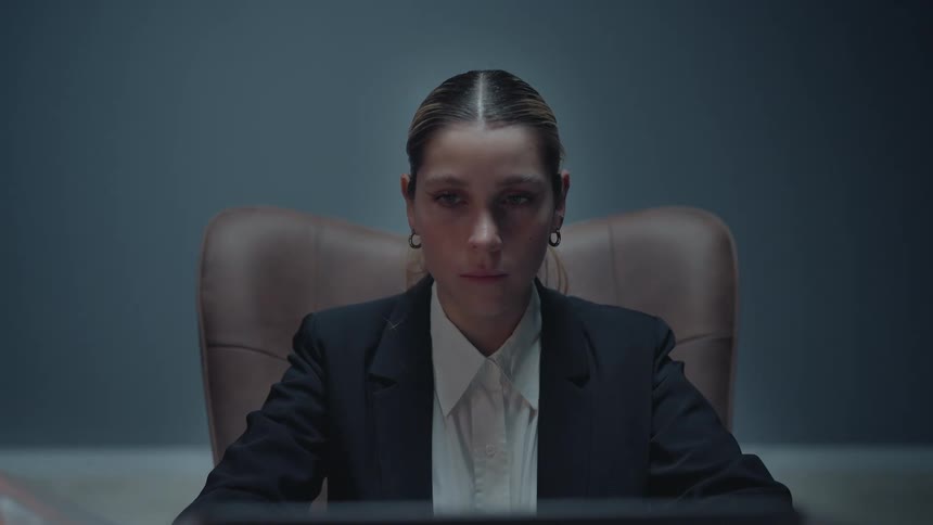 A serious businesswoman in a dimly lit office focuses intensely on her laptop as the camera slowly zooms in