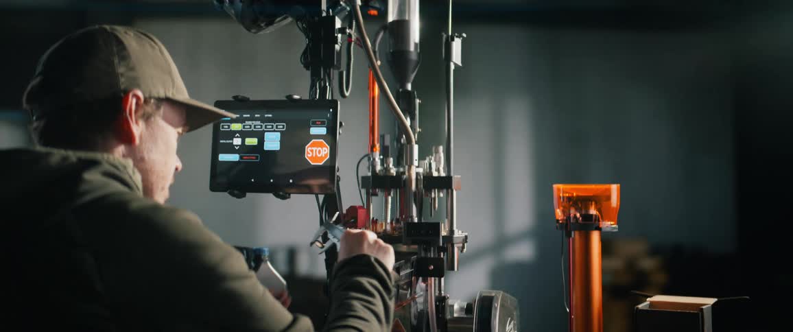A man in a workshop carefully operates an automated ammunition reloading press, using a tablet and calipers to ensure precision