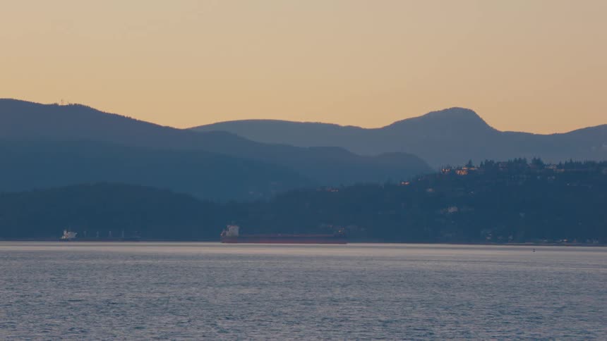 A peaceful seascape shows a cargo ship floating near silhouetted mountains under a warm sunset sky