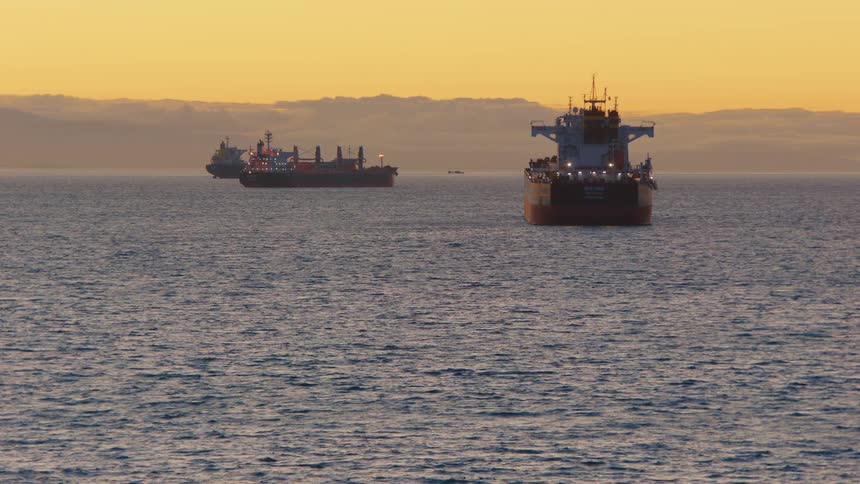 Large cargo ships are anchored in the calm ocean against a beautiful golden sunset sky