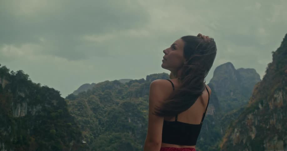 A serene woman with her hair blowing in the wind gazes up at the sky in front of a stunning mountain range