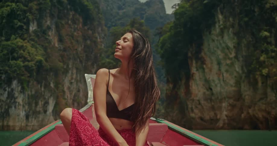 A tranquil woman with eyes closed enjoys the warmth of the sun while sitting on the bow of a boat, gliding through a stunning tropical canyon lake