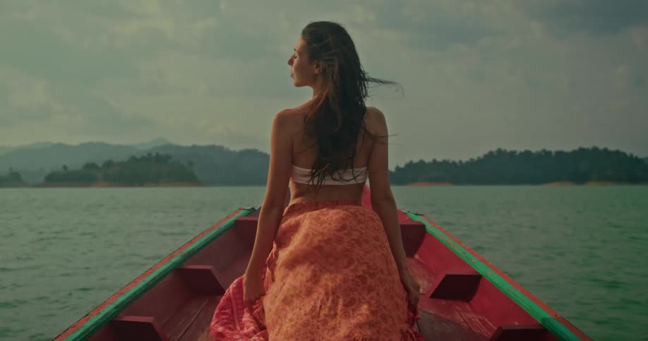 A woman in a colorful skirt sits on the bow of a boat as it glides across a serene lake, with the wind blowing through her hair
