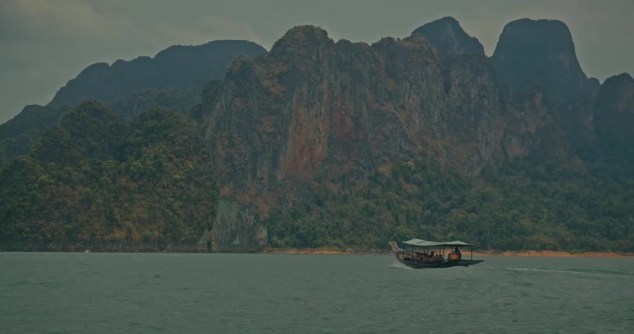 A traditional long-tail boat full of tourists cruises across a vast lake surrounded by dramatic jungle-covered mountains