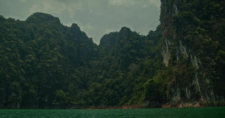 Slow tilt-down from a cloudy sky revealing the majestic, forest-covered mountains surrounding a tranquil green lake
