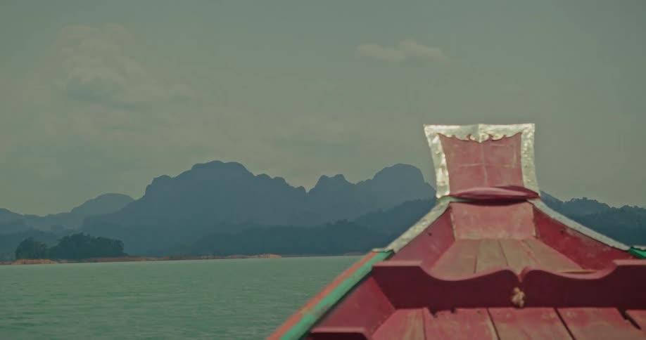 A scenic view from the bow of a traditional wooden boat sailing across a vast lake towards distant, hazy mountains under an overcast sky