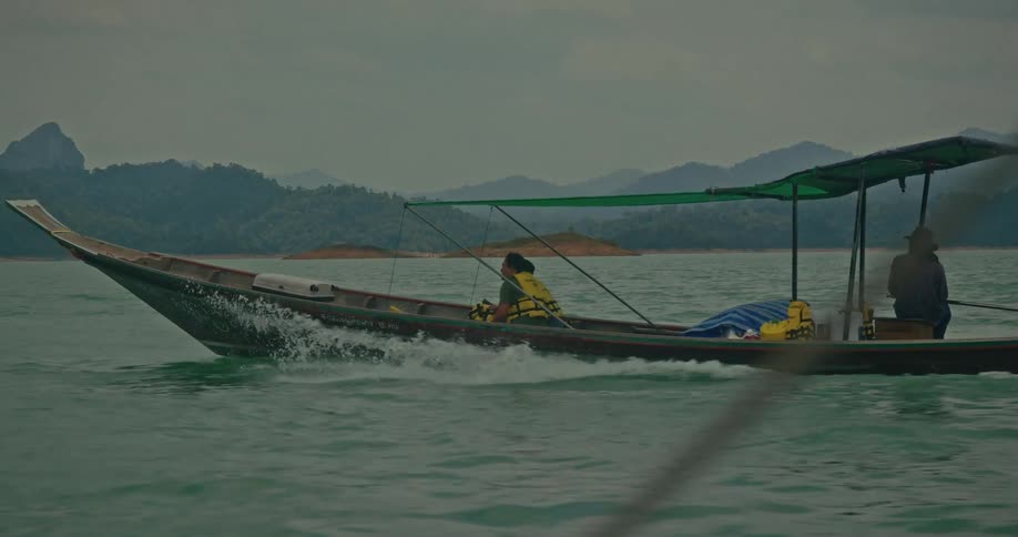 A traditional longtail boat ferries passengers across a vast lake surrounded by mountains on a cloudy day, captured in slow motion