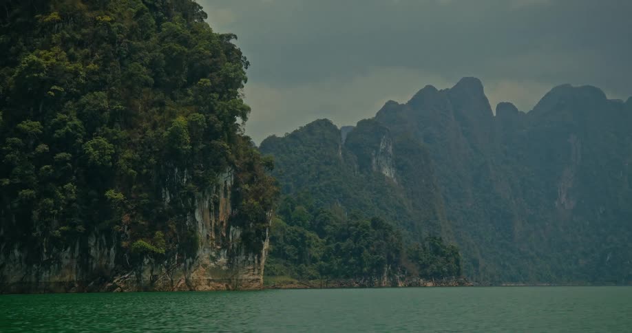 A serene view of a calm lake surrounded by towering, jungle-covered limestone mountains under a cloudy sky