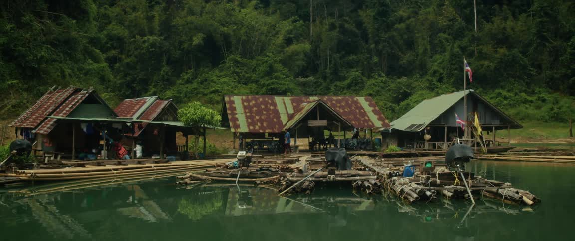 A rustic floating village on bamboo rafts sits on a calm river in the dense Thai jungle