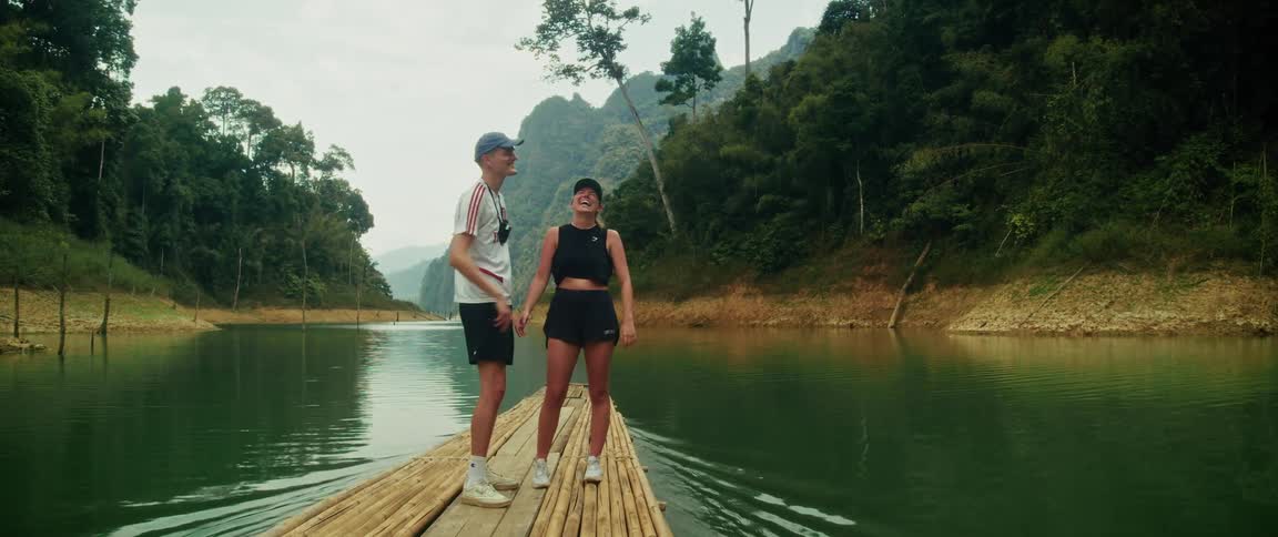 A young, happy couple enjoys the scenic jungle views while floating down a tranquil lake on a bamboo raft