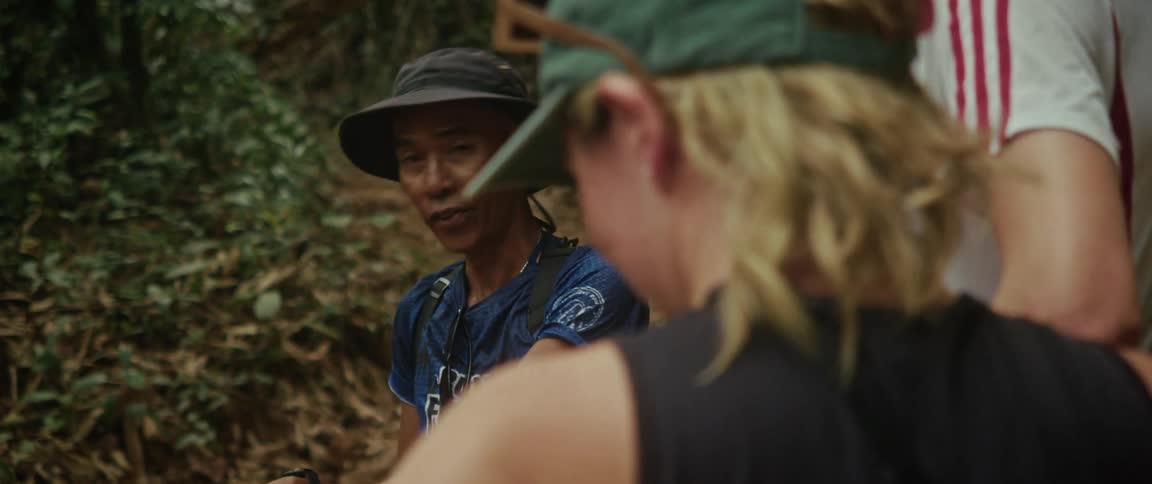 A friendly Asian tour guide in a hiking hat explains the trail to a group of tourists deep in a lush, green jungle