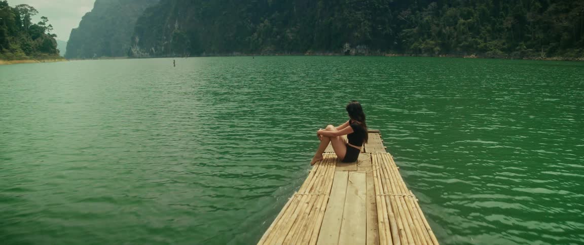 A serene woman enjoys a peaceful journey on a bamboo raft, gliding across a vast emerald lake nestled among lush green mountains