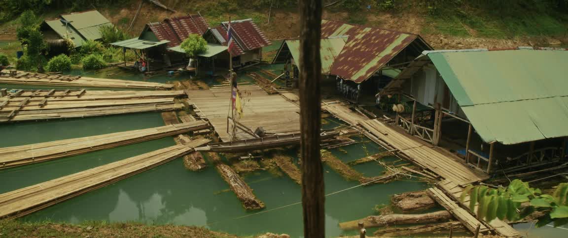 A high-angle dolly shot reveals a rustic floating village with wooden huts and people on the docks in a lush, green jungle