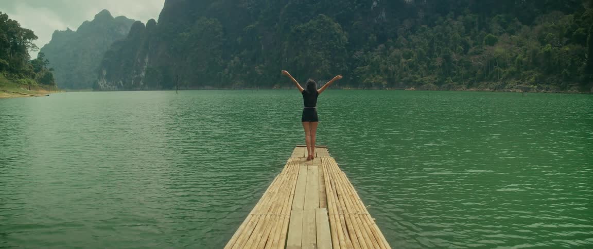 A carefree woman on a bamboo raft embraces the stunning natural landscape of a green mountain lake, raising her arms in freedom