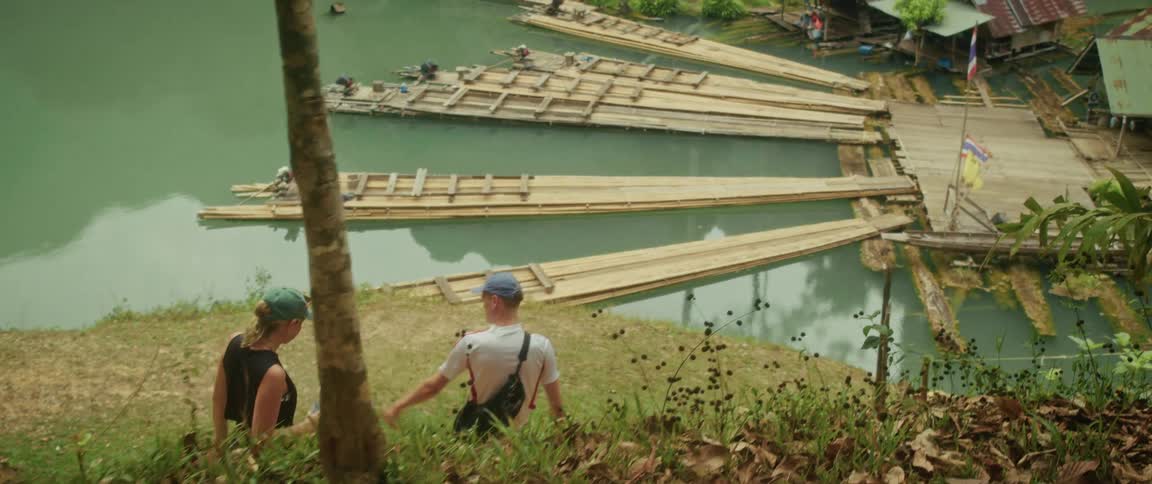 A traveling couple hikes down a trail overlooking a tranquil river with bamboo rafts and a floating village in Thailand