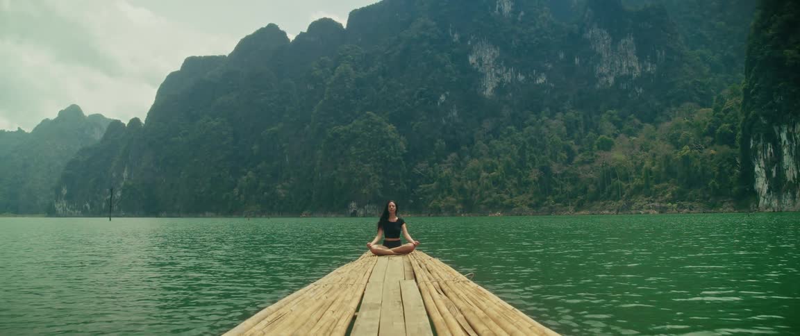 A woman meditates peacefully on a bamboo raft floating on a tranquil green lake surrounded by majestic mountains