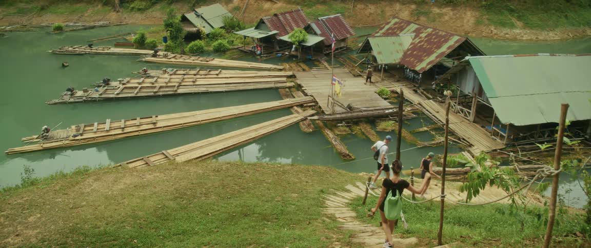 A group of tourists walks down a path towards a floating village with bamboo rafts on a lake in a lush jungle