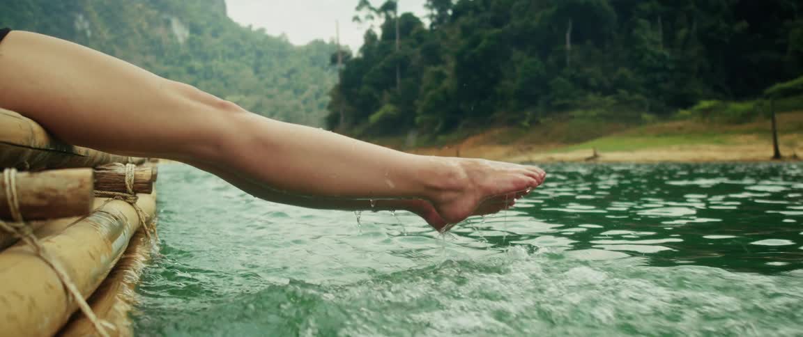 A woman's legs and feet splash playfully in the green lake water from the side of a bamboo raft in slow motion