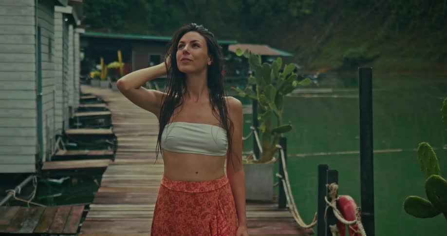 A serene woman with wet hair walks along a wooden pier at a tropical lake resort during a light rain shower